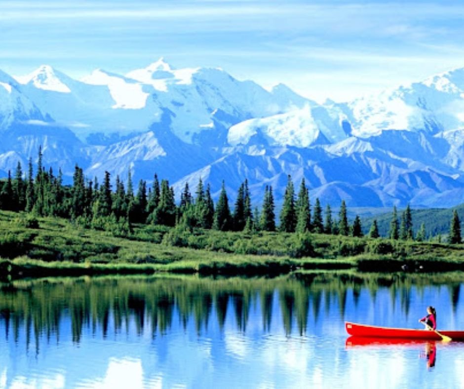 Person in a red canoe on a serene lake with snowy mountains and forest in the background.