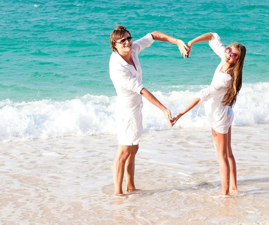 A couple in white dress making a heart shape with their arms on a beach.