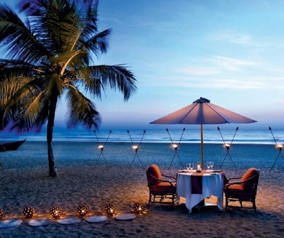 Romantic beach dinner setup with lit torches, umbrella, and a palm tree at twilight.