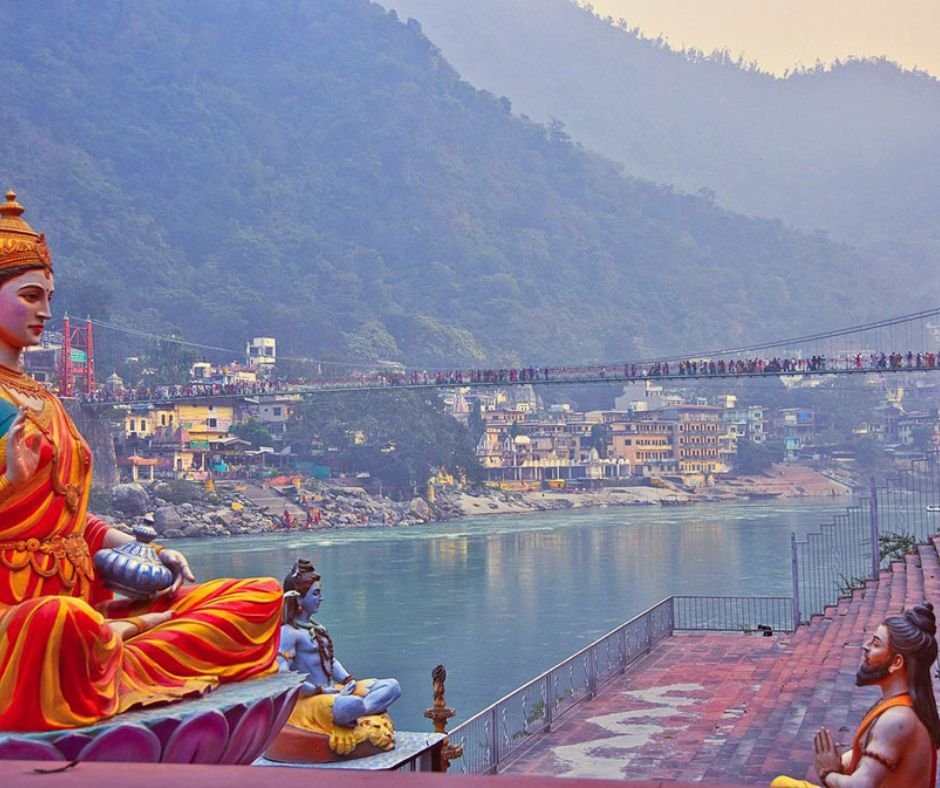 A colorful riverside with statues in the foreground and a crowded suspension bridge in the background.