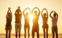 Shadow of six people forming heart shapes with their hands on a beach at sunset.