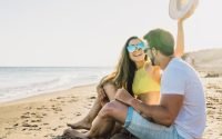 A couple sitting on the beach with a man holding a hat in the air.