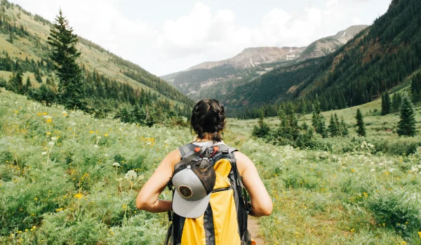 A person with a backpack walks through a lush green valley surrounded by mountains and wildflowers. The sky is partly cloudy. The person is wearing a cap attached to the backpack.